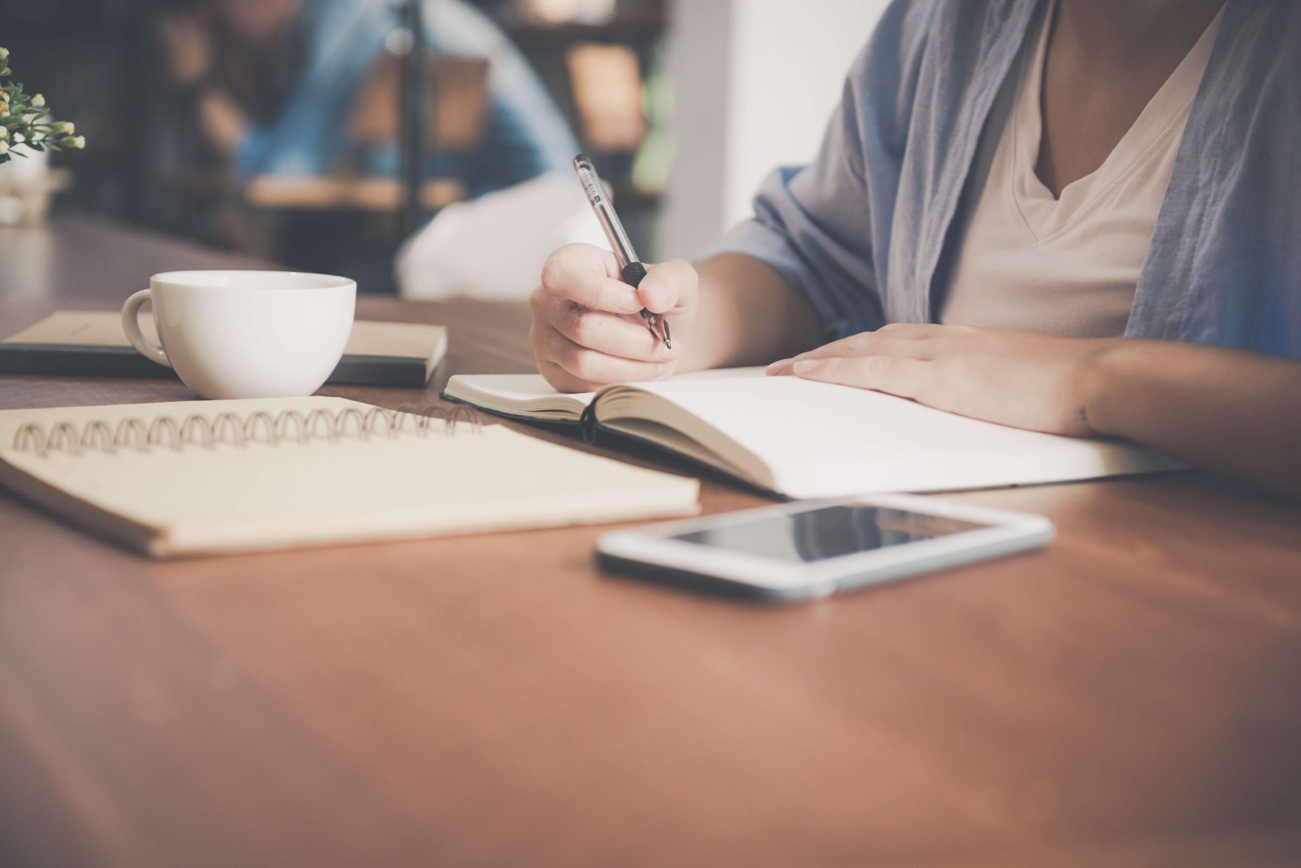 Consulta extrato GPS: Pessoa escrevendo em caderno que está sobre a mesa. Sobre a mesa também estão: outro caderno aberto, um celular e uma xícara.