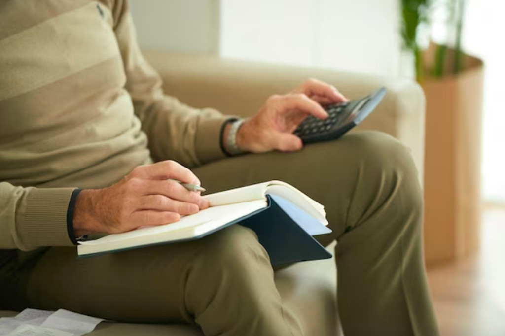 Homem com mãos levemente enrugadas sentado em sofá, usando uma calculadora sobre um joelho e escrevendo num caderno que está sobre o outro joelho. Simbolizando os cálculos do fator previdenciário e aposentandoria