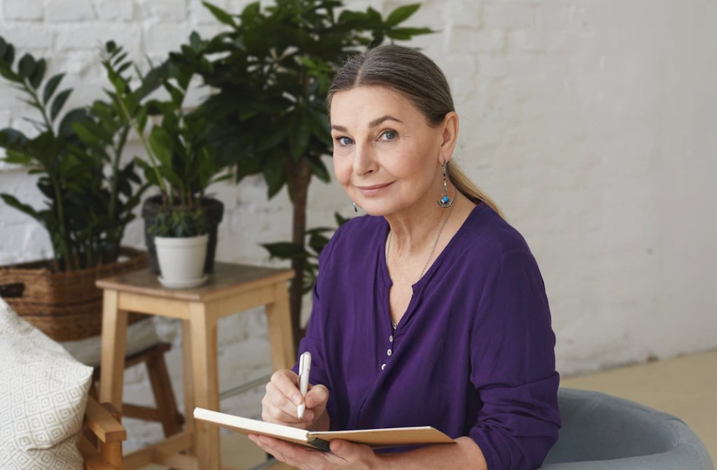 Dentro de uma sala de estar com plantas, uma senhora está sentada escrevendo num caderno, simbolizando um plano de aposentadoria