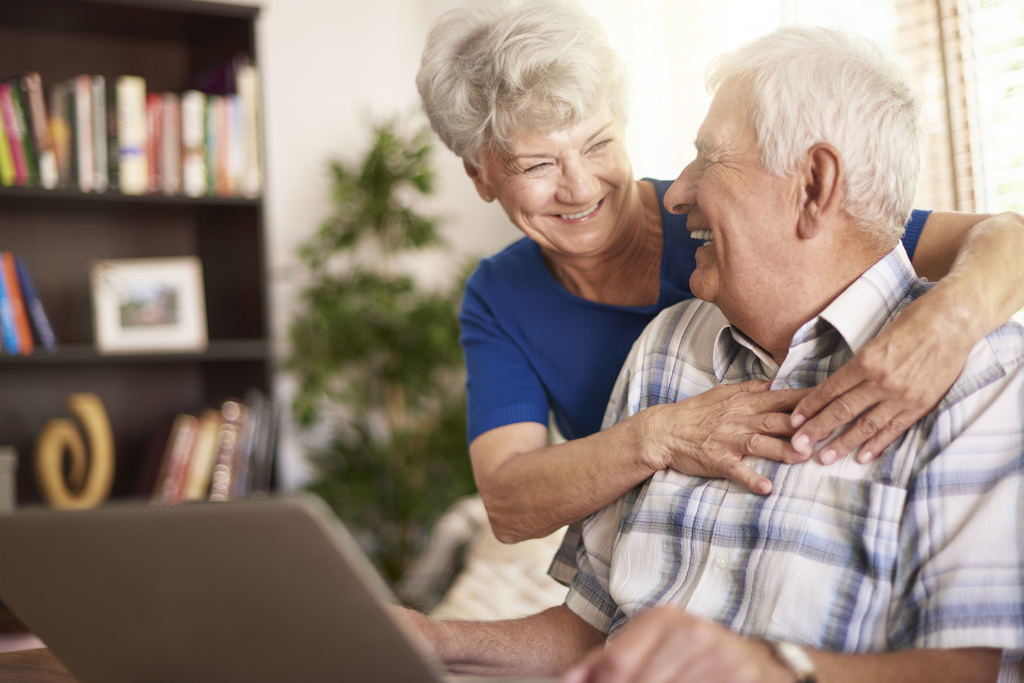 Simbolizando um bom plano de aposentadoria, um casal de idosos está sorrindo e se abraçando, enquanto ele usa um notebook. Ao fundo da sala está uma estante com livros, um porta-retrato e uma pequena estátua abstrato