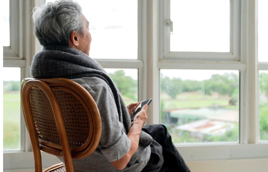 Pessoa idosa sentada numa cadeira olhando pela janela, segurando um smartphone