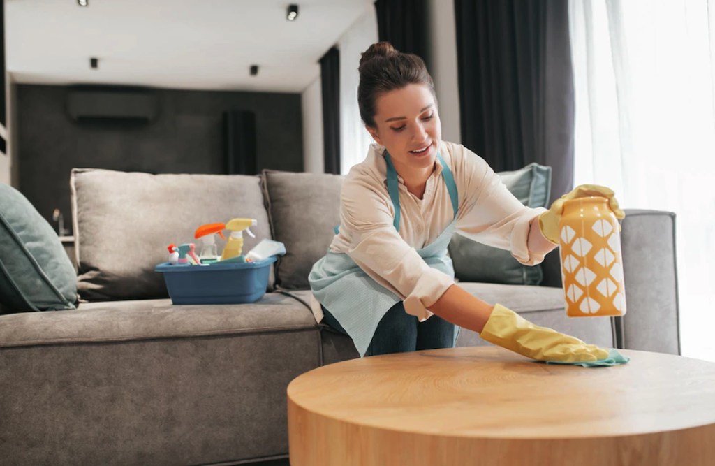 Simbolizando aposentadoria para doméstica do lar: uma mulher em sala de estar limpando mesinha de centro com uma flanela. A mulher está usando avental e luvas e sentada em um sofá, sobre o sofá está uma caixa com produtos de limpeza.