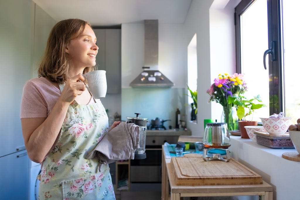 Mulher segurando uma xícara de café e um pano, usando um avental, sorrindo olhando pela janela da cozinha, simbolizando aposentadoria para doméstica do lar