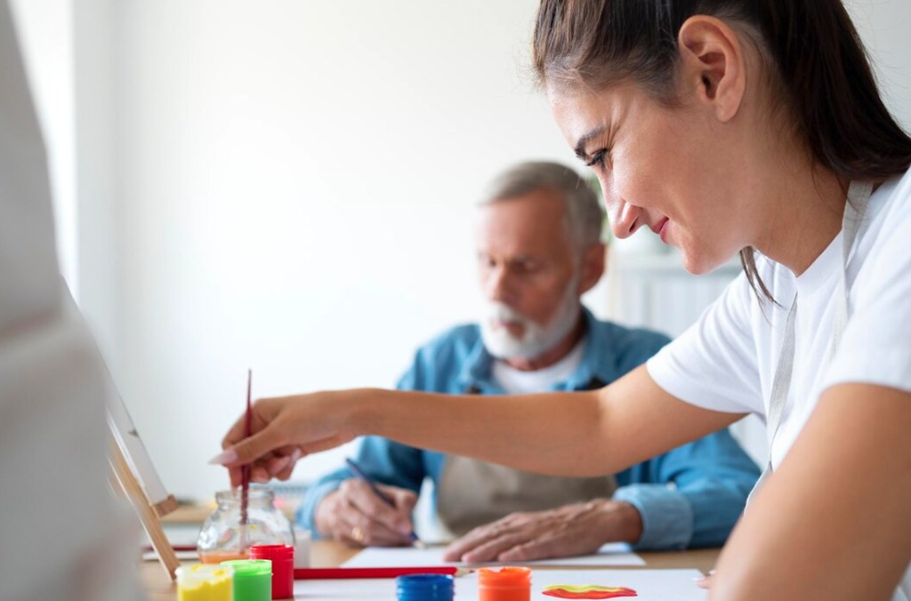 Mulher com camiseta branca fazendo pintura com tintas sorrindo, e um senhor no fundo usando um lápis num papel, simbolizando aposentadoria por autismo