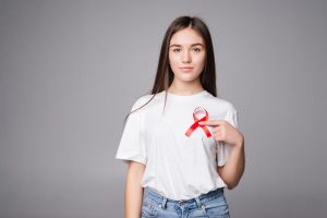 Mulher em fundo neutro, cinza, com camiseta branca, segurando um laço vermelho sobre o lado esquerdo do peito, na altura do coração. O laço simboliza a luta contra a AIDS,
