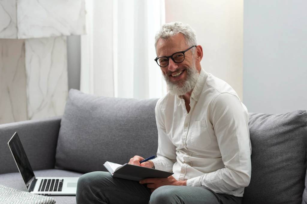 Senhor de cabelo e barba brancos, com óculos de grau, sentado num sofá com notebook no assento ao lado, sorrindo e escrevendo numa agenda. Simbolizando estabilidade pré-aposentadoria