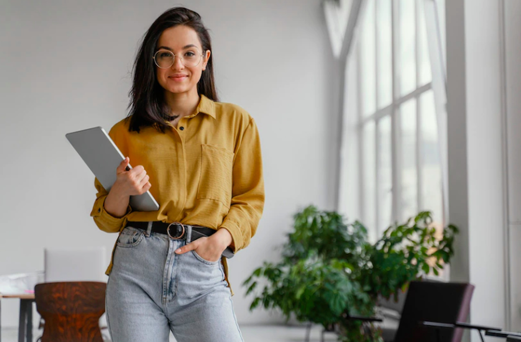 Estagiária de óculos de grau, sorrindo com uma mão no bolso da calça jeans e outra mão segurando um tablet