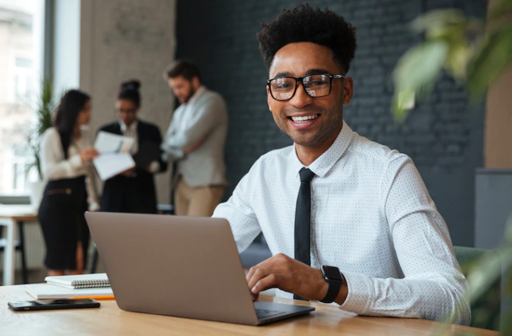 Jovem de camisa social e gravada, de óculos de grau e um largo sorrindo olhando para a câmera, enquanto usa o teclado de um notebook em um ambiente corporativo moderno, simbolizando um estagiário