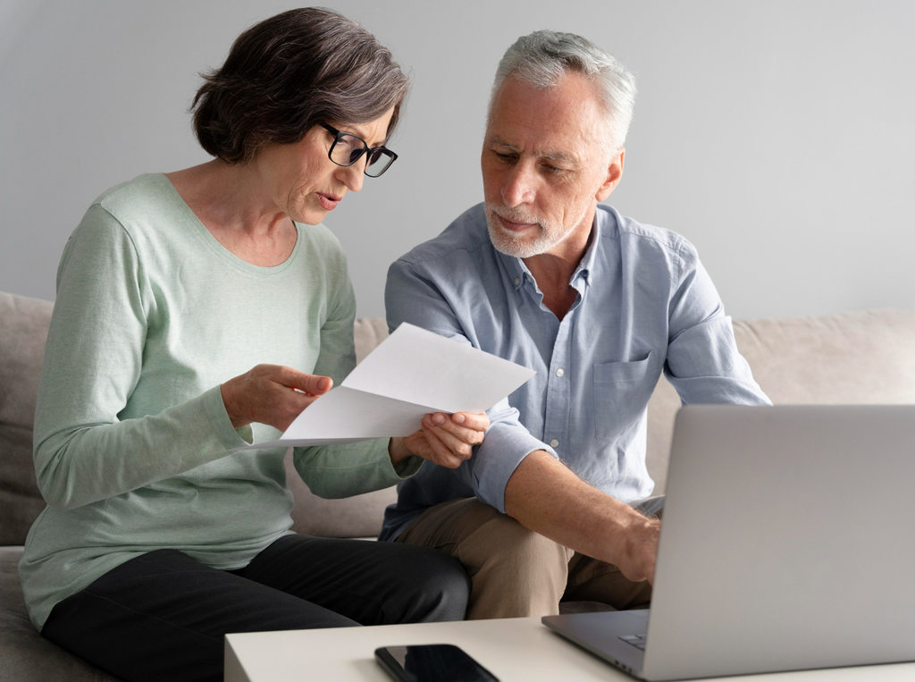 Casal de idosos analisando um documento em papel, simbolizando como aumentar o valor da aposentadoria. Na frente deles há um celular e um notebook