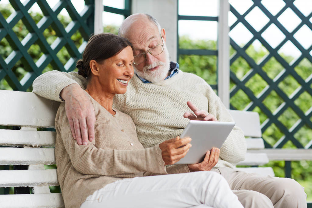 Casal de idosos. Ela sorrindo, ele interagindo e ambos olhando para um tablet, simbolizando como dar entrada na aposentadoria INSS