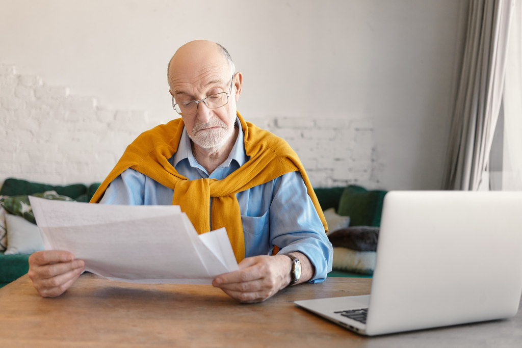Simbolizando dicas de como aumentar o valor da aposentadoria: Senhor analisando documentos em papel, na frente dele está um notebook sobre a mesa na qual seus braços estão apoiados