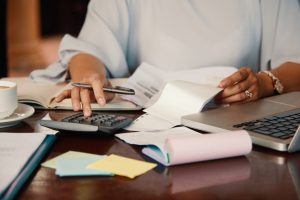 Pessoa usando calculadora e analisando documentos que estão sobre a mesa de escritório, junto com um notebook
