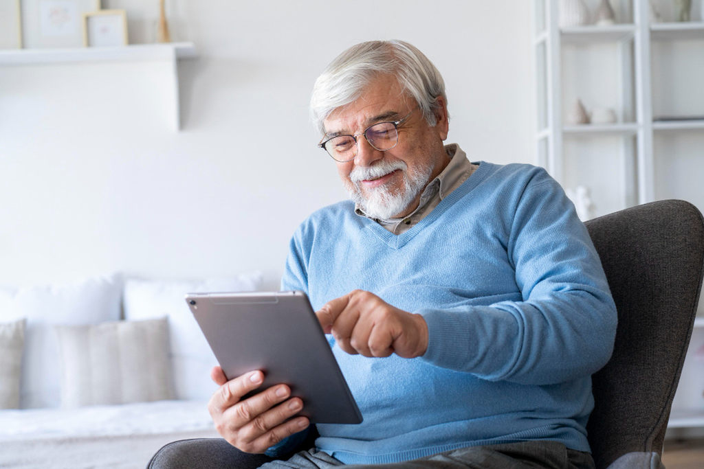 Aposentado sorrindo de cabelo e barba brancos, e óculos de grau, olhando para tablet, simbolizando ganhos após revisão
