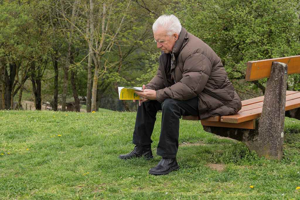 Senhor de cabelos brancos e óculos de grau lendo um livro sentado em banco de um parque com grama e árvores, simbolizando revisão da vida toda