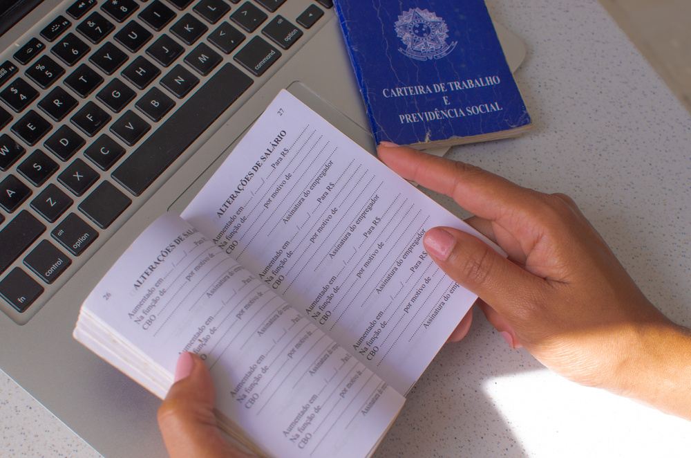 Pessoa com notebook e carteira de trabalho nas mãos, possivelmente pesquisando sobre os indicadores CNIS