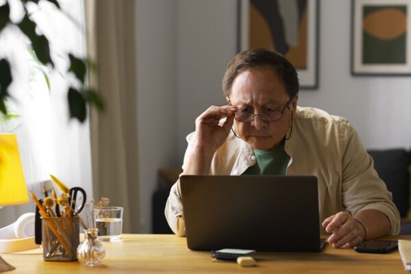Homem idoso com expressão de dúvida e preocupação usando óculos e olhando para a tela de um notebook enquanto tenta simular sua aposentadoria por idade em 2026.